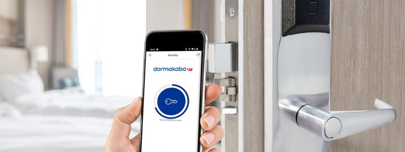 Verkada Installers A hand holds a smartphone displaying a digital key app near a modern electronic door lock, highlighting the seamless integration of Jendy Physical Security solutions, with a blurred hotel room visible in the background.