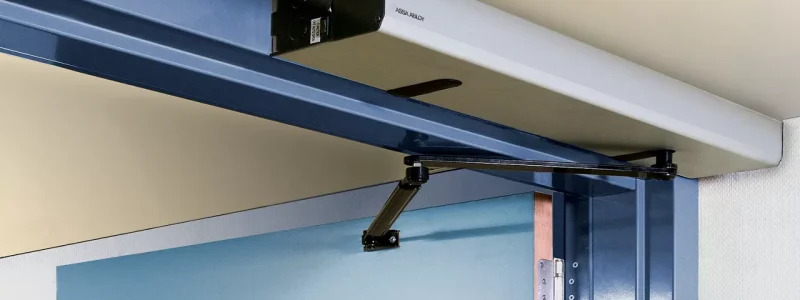 Verkada Installers Automatic door closer mechanism mounted above a blue swinging door, attached to a metal frame at the top corner, with a black arm connecting the device to the door—expertly installed by Verkada Installers Jendy Physical Security.