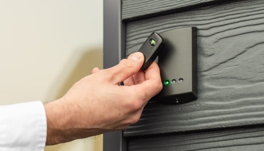 Verkada Installers A person’s hand holds a key fob up to an electronic card reader mounted on a gray textured wall, possibly installed by Verkada Installers Jendy Physical Security, to unlock a secure door.