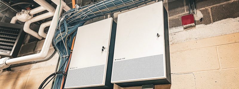 Verkada Installers Two white electrical or network cabinets are mounted on a beige wall in an industrial setting, with cables running above and beside them. A red fire alarm and exposed pipes are also visible. The Jendy logo appears in the lower right corner.