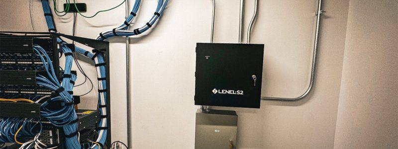 Verkada Installers A tidy server room wall with neatly arranged blue cables, electrical conduits, a LENEL S2 control panel, a smaller gray panel, and a Jendy Technologies logo—expertly organized by Verkada Installation security system Verkada installers.