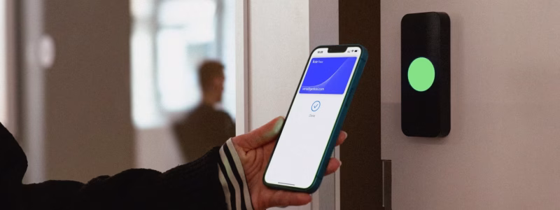 Verkada Installers A person holds a smartphone with a digital access pass near a wall-mounted electronic reader displaying a green light, indicating successful access. Verkada Installers Jendy Physical Security ensure seamless integration in the background.