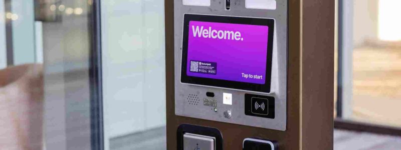 Verkada Installers A kiosk in a building lobby displays a purple screen that says “Welcome. Tap to start.” Installed by Verkada Installers Jendy, it features buttons, a card reader, speaker, and an accessibility button to open the door for enhanced physical security.