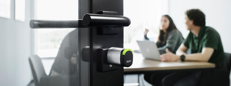 Verkada Installers A close-up of a modern, electronic door lock by Verkada Installers on a glass door, with two people at a table in a bright office in the background, one using a laptop—highlighting smart solutions for physical security.